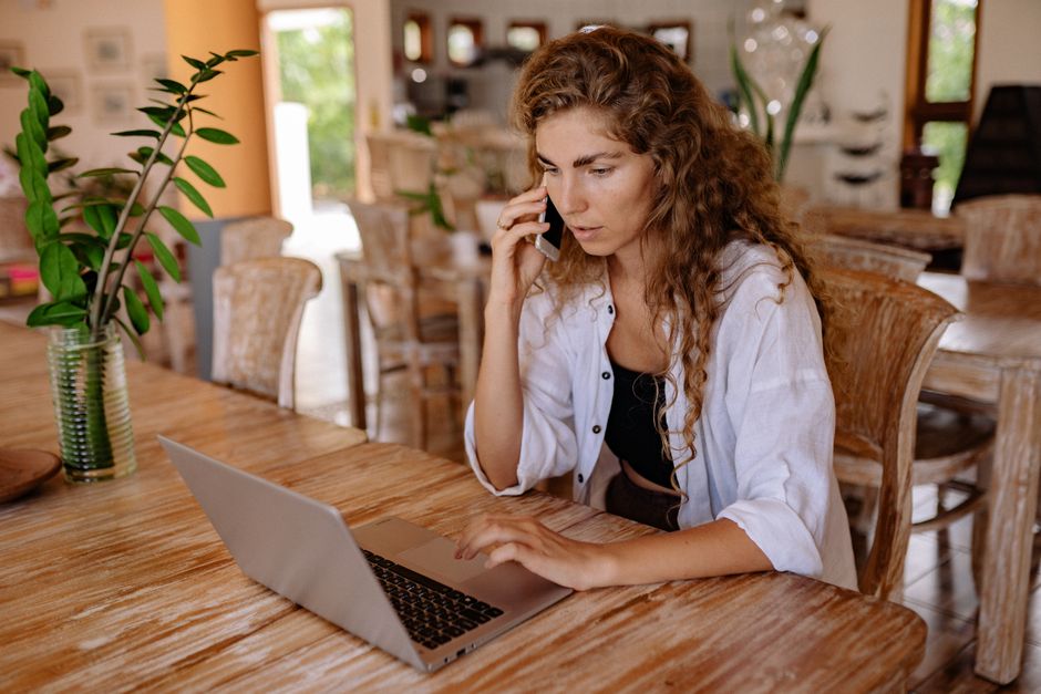 A woman in a bright room using a laptop and phone, blending work with home comfort.